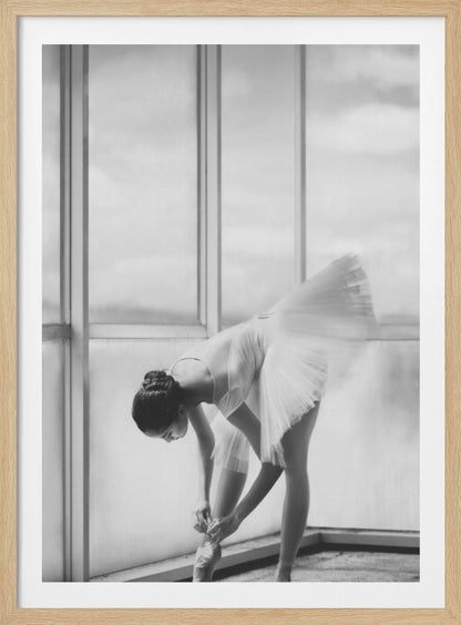 A black and white photograph of a ballerina in a white tutu, bending over to tie her pointe shoe in front of a large, bright window. The motion of her tutu is captured in a graceful blur. Print