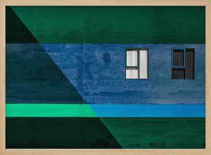 A framed abstract architectural photograph featuring a color-blocked facade. A central horizontal band of textured blue wall contains two small, distinct windows. This is flanked by upper and lower sections of dark green. A large, dark diagonal shape overlays the left side of the composition, creating sharp geometric lines. Decor