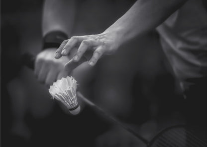 A dramatic black and white close-up photograph of a badminton player preparing to serve. The focus is on the player's hands, with one hand delicately positioned above a shuttlecock which is balanced on the strings of a racquet. Artwork