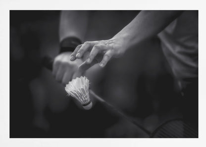 A dramatic black and white close-up photograph of a badminton player preparing to serve. The focus is on the player's hands, with one hand delicately positioned above a shuttlecock which is balanced on the strings of a racquet. Artwork