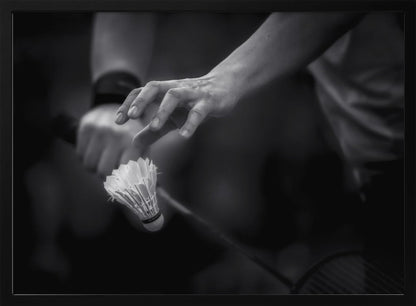 A dramatic black and white close-up photograph of a badminton player preparing to serve. The focus is on the player's hands, with one hand delicately positioned above a shuttlecock which is balanced on the strings of a racquet. Artwork