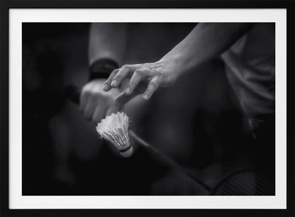 A dramatic black and white close-up photograph of a badminton player preparing to serve. The focus is on the player's hands, with one hand delicately positioned above a shuttlecock which is balanced on the strings of a racquet. Artwork