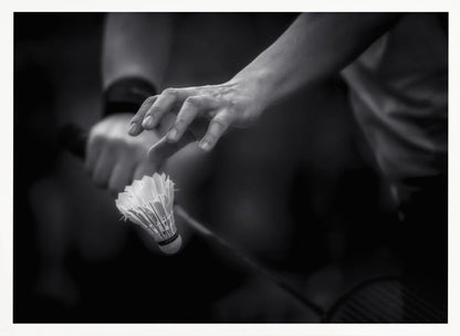 A dramatic black and white close-up photograph of a badminton player preparing to serve. The focus is on the player's hands, with one hand delicately positioned above a shuttlecock which is balanced on the strings of a racquet. Artwork