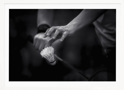A dramatic black and white close-up photograph of a badminton player preparing to serve. The focus is on the player's hands, with one hand delicately positioned above a shuttlecock which is balanced on the strings of a racquet. Artwork
