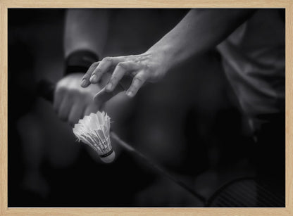 A dramatic black and white close-up photograph of a badminton player preparing to serve. The focus is on the player's hands, with one hand delicately positioned above a shuttlecock which is balanced on the strings of a racquet. Artwork