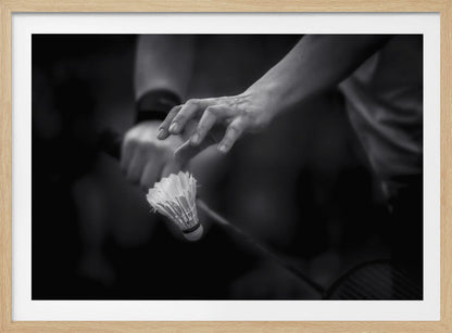 A dramatic black and white close-up photograph of a badminton player preparing to serve. The focus is on the player's hands, with one hand delicately positioned above a shuttlecock which is balanced on the strings of a racquet. Artwork
