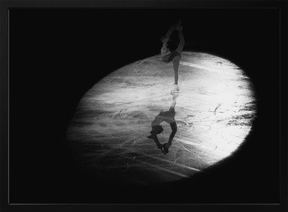 A dramatic black and white framed photograph of a female figure skater performing on ice. She is illuminated by a single spotlight, holding a graceful standing split pose, with her reflection visible on the ice below. Poster