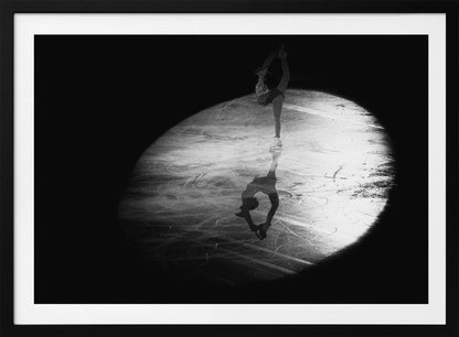 A dramatic black and white framed photograph of a female figure skater performing on ice. She is illuminated by a single spotlight, holding a graceful standing split pose, with her reflection visible on the ice below. Poster