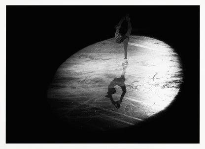 A dramatic black and white framed photograph of a female figure skater performing on ice. She is illuminated by a single spotlight, holding a graceful standing split pose, with her reflection visible on the ice below. Poster