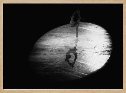 A dramatic black and white framed photograph of a female figure skater performing on ice. She is illuminated by a single spotlight, holding a graceful standing split pose, with her reflection visible on the ice below. Poster