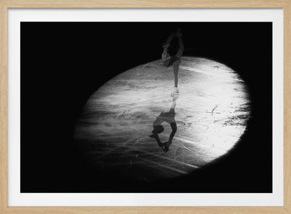 A dramatic black and white framed photograph of a female figure skater performing on ice. She is illuminated by a single spotlight, holding a graceful standing split pose, with her reflection visible on the ice below. Poster