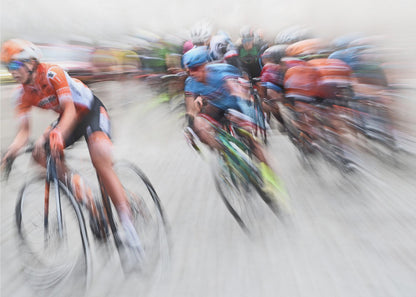 An action photograph of a professional cycling race captured with a prominent motion blur effect that conveys a strong sense of speed. A cyclist in an orange and black uniform is in the foreground, leaning into a turn, while a group of other riders in blue and multi-colored jerseys follows closely behind, their forms streaked and blurred. The entire scene is encased in a brushed silver frame. Poster
