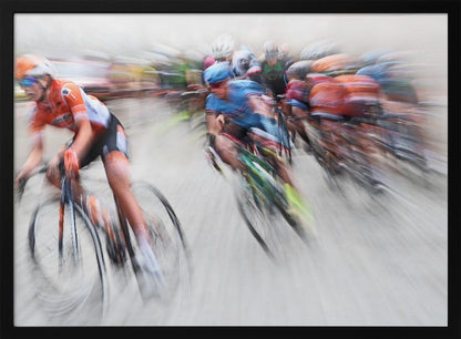 An action photograph of a professional cycling race captured with a prominent motion blur effect that conveys a strong sense of speed. A cyclist in an orange and black uniform is in the foreground, leaning into a turn, while a group of other riders in blue and multi-colored jerseys follows closely behind, their forms streaked and blurred. The entire scene is encased in a brushed silver frame. Poster
