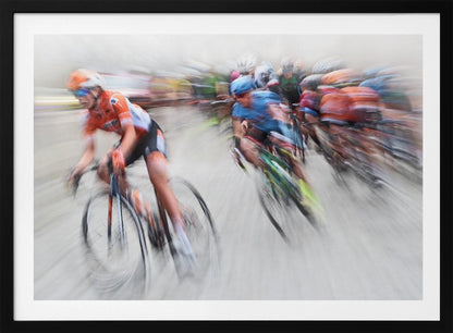 An action photograph of a professional cycling race captured with a prominent motion blur effect that conveys a strong sense of speed. A cyclist in an orange and black uniform is in the foreground, leaning into a turn, while a group of other riders in blue and multi-colored jerseys follows closely behind, their forms streaked and blurred. The entire scene is encased in a brushed silver frame. Poster