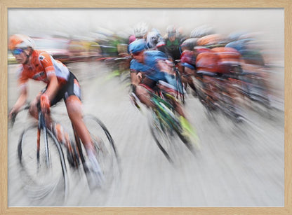 An action photograph of a professional cycling race captured with a prominent motion blur effect that conveys a strong sense of speed. A cyclist in an orange and black uniform is in the foreground, leaning into a turn, while a group of other riders in blue and multi-colored jerseys follows closely behind, their forms streaked and blurred. The entire scene is encased in a brushed silver frame. Poster