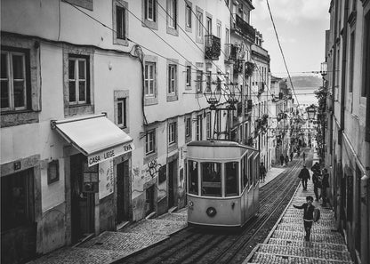 A black and white photograph of a vintage tram on a steep, narrow cobblestone street in an old European city. The street is lined with historic buildings and people are walking alongside the tracks. In the distance, a body of water is visible. Poster
