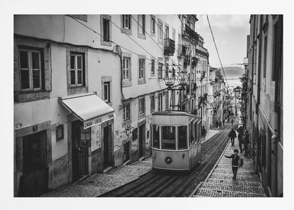 A black and white photograph of a vintage tram on a steep, narrow cobblestone street in an old European city. The street is lined with historic buildings and people are walking alongside the tracks. In the distance, a body of water is visible. Poster