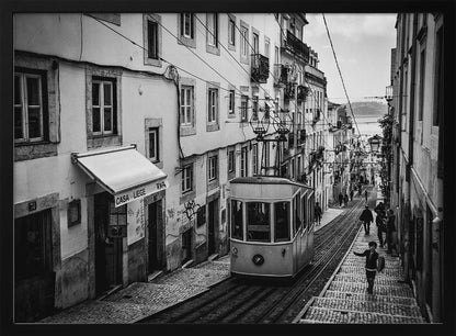 A black and white photograph of a vintage tram on a steep, narrow cobblestone street in an old European city. The street is lined with historic buildings and people are walking alongside the tracks. In the distance, a body of water is visible. Poster