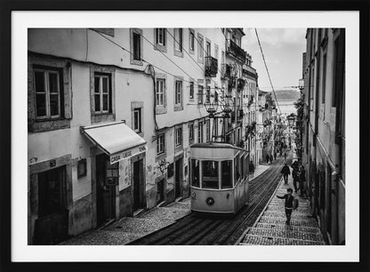A black and white photograph of a vintage tram on a steep, narrow cobblestone street in an old European city. The street is lined with historic buildings and people are walking alongside the tracks. In the distance, a body of water is visible. Poster