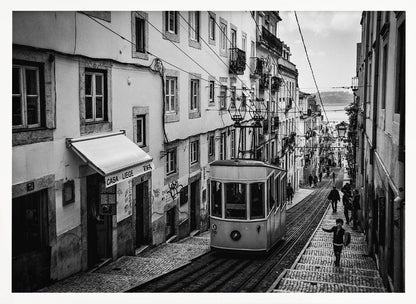 A black and white photograph of a vintage tram on a steep, narrow cobblestone street in an old European city. The street is lined with historic buildings and people are walking alongside the tracks. In the distance, a body of water is visible. Poster