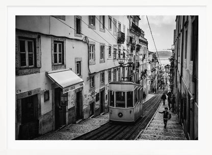 A black and white photograph of a vintage tram on a steep, narrow cobblestone street in an old European city. The street is lined with historic buildings and people are walking alongside the tracks. In the distance, a body of water is visible. Poster