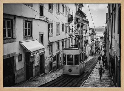 A black and white photograph of a vintage tram on a steep, narrow cobblestone street in an old European city. The street is lined with historic buildings and people are walking alongside the tracks. In the distance, a body of water is visible. Poster