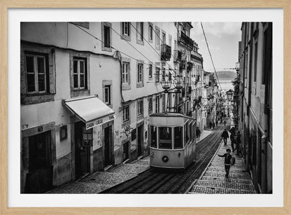 A black and white photograph of a vintage tram on a steep, narrow cobblestone street in an old European city. The street is lined with historic buildings and people are walking alongside the tracks. In the distance, a body of water is visible. Poster