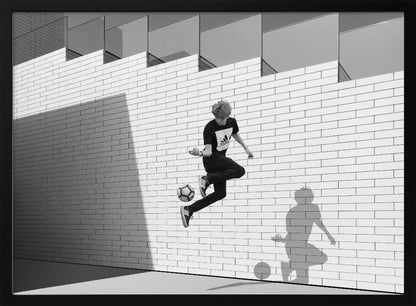 A framed, black and white photograph of a young person in mid-air, skillfully juggling a soccer ball with their foot against a clean, white brick wall. The person's shadow is cast sharply on the wall, mirroring the action, under a modern building with angled windows. Wall Art