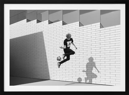 A framed, black and white photograph of a young person in mid-air, skillfully juggling a soccer ball with their foot against a clean, white brick wall. The person's shadow is cast sharply on the wall, mirroring the action, under a modern building with angled windows. Wall Art