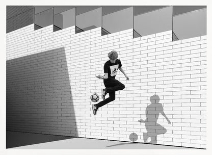 A framed, black and white photograph of a young person in mid-air, skillfully juggling a soccer ball with their foot against a clean, white brick wall. The person's shadow is cast sharply on the wall, mirroring the action, under a modern building with angled windows. Wall Art