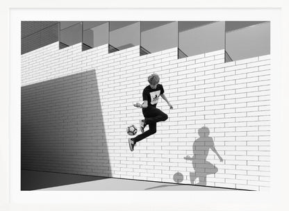 A framed, black and white photograph of a young person in mid-air, skillfully juggling a soccer ball with their foot against a clean, white brick wall. The person's shadow is cast sharply on the wall, mirroring the action, under a modern building with angled windows. Wall Art