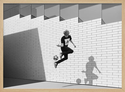 A framed, black and white photograph of a young person in mid-air, skillfully juggling a soccer ball with their foot against a clean, white brick wall. The person's shadow is cast sharply on the wall, mirroring the action, under a modern building with angled windows. Wall Art