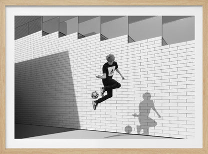 A framed, black and white photograph of a young person in mid-air, skillfully juggling a soccer ball with their foot against a clean, white brick wall. The person's shadow is cast sharply on the wall, mirroring the action, under a modern building with angled windows. Wall Art