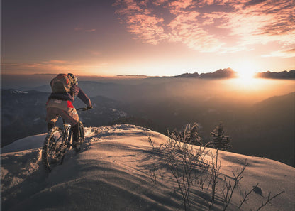 A mountain biker seen from behind pauses on a snowy mountain summit, overlooking a vast, misty mountain range as the sun sets, casting a warm orange and pink glow across the sky and snow. Artwork