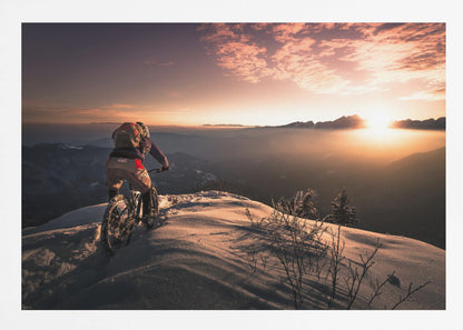 A mountain biker seen from behind pauses on a snowy mountain summit, overlooking a vast, misty mountain range as the sun sets, casting a warm orange and pink glow across the sky and snow. Artwork