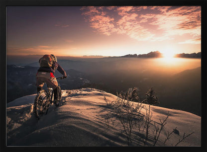 A mountain biker seen from behind pauses on a snowy mountain summit, overlooking a vast, misty mountain range as the sun sets, casting a warm orange and pink glow across the sky and snow. Artwork