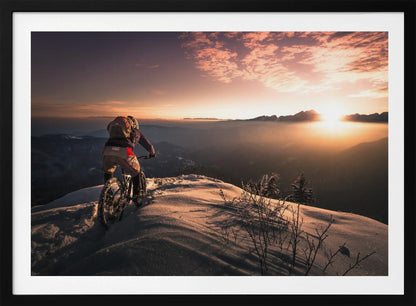 A mountain biker seen from behind pauses on a snowy mountain summit, overlooking a vast, misty mountain range as the sun sets, casting a warm orange and pink glow across the sky and snow. Artwork