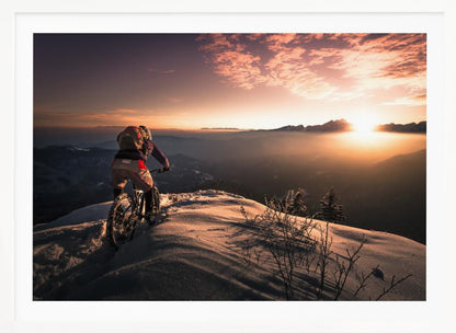 A mountain biker seen from behind pauses on a snowy mountain summit, overlooking a vast, misty mountain range as the sun sets, casting a warm orange and pink glow across the sky and snow. Artwork