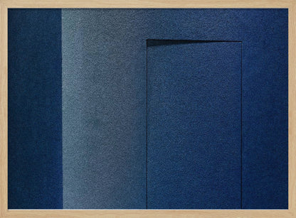 An abstract geometric artwork featuring textured panels in various shades of deep and dusty blue. The composition is divided into vertical sections, with a central rectangular shape that appears slightly recessed, creating a strong shadow line at its top edge. The piece is enclosed in a simple, brushed silver frame. Wall Art