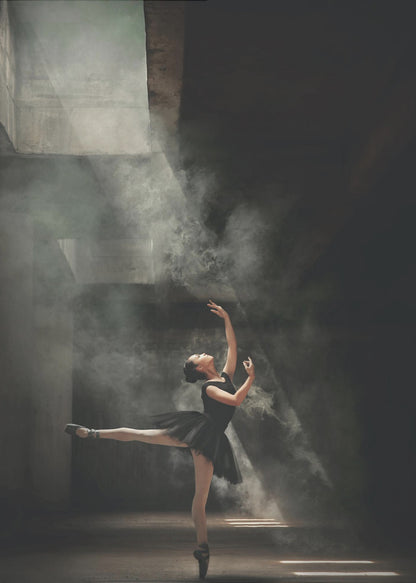 A ballerina in a black tutu gracefully performs an arabesque in a vast, dark, industrial space filled with smoke, dramatically illuminated by shafts of light from above. Poster