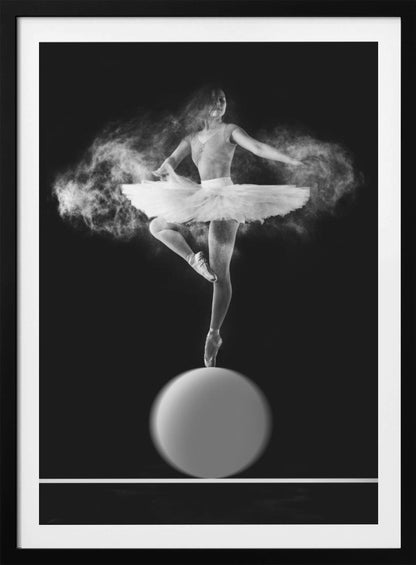 A dramatic black and white photograph of a ballerina balancing on one pointe shoe on top of a large sphere. She wears a white tutu and is surrounded by a cloud of white powder, capturing a moment of explosive, graceful movement against a solid black background. Wall Art