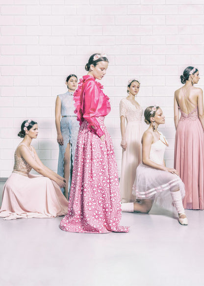 A studio photograph of six young women in elegant pastel gowns posing against a white brick wall. The central figure wears a vibrant pink ruffled blouse and a patterned maxi skirt, while the others are dressed in light pink, white, and pale blue dresses, some with ballet-inspired elements. They all have flowers in their hair, creating a soft, high-fashion aesthetic. Print