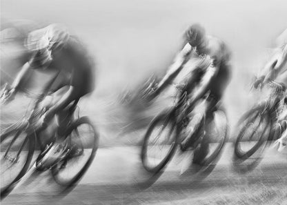 A dynamic black and white action photograph of three cyclists racing, captured with a slow shutter speed to create a dramatic motion blur effect. The riders and their bikes are blurred into streaks of light and shadow, conveying a powerful sense of speed and intensity. The image is presented within a silver-toned frame. Poster