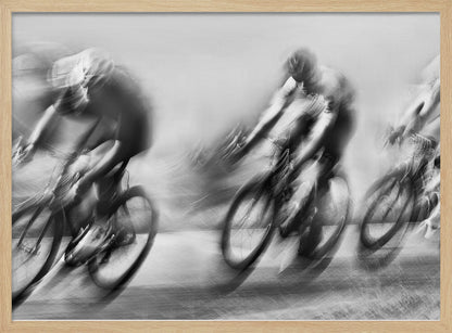 A dynamic black and white action photograph of three cyclists racing, captured with a slow shutter speed to create a dramatic motion blur effect. The riders and their bikes are blurred into streaks of light and shadow, conveying a powerful sense of speed and intensity. The image is presented within a silver-toned frame. Poster