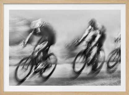 A dynamic black and white action photograph of three cyclists racing, captured with a slow shutter speed to create a dramatic motion blur effect. The riders and their bikes are blurred into streaks of light and shadow, conveying a powerful sense of speed and intensity. The image is presented within a silver-toned frame. Poster