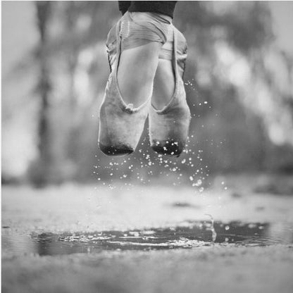 A close-up, black and white photograph of a ballerina's feet in worn pointe shoes, captured in mid-air above a puddle, with water splashing upwards. The motion is frozen, highlighting the contrast between grace and a raw outdoor element. The image is presented in a light wood frame. Poster