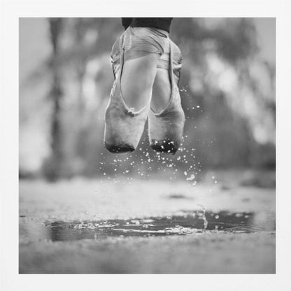 A close-up, black and white photograph of a ballerina's feet in worn pointe shoes, captured in mid-air above a puddle, with water splashing upwards. The motion is frozen, highlighting the contrast between grace and a raw outdoor element. The image is presented in a light wood frame. Poster