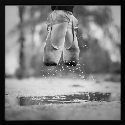 A close-up, black and white photograph of a ballerina's feet in worn pointe shoes, captured in mid-air above a puddle, with water splashing upwards. The motion is frozen, highlighting the contrast between grace and a raw outdoor element. The image is presented in a light wood frame. Poster