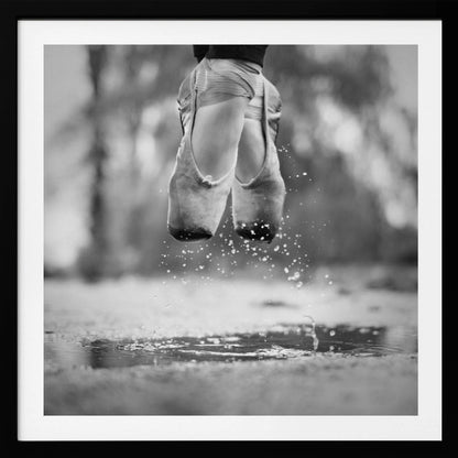 A close-up, black and white photograph of a ballerina's feet in worn pointe shoes, captured in mid-air above a puddle, with water splashing upwards. The motion is frozen, highlighting the contrast between grace and a raw outdoor element. The image is presented in a light wood frame. Poster