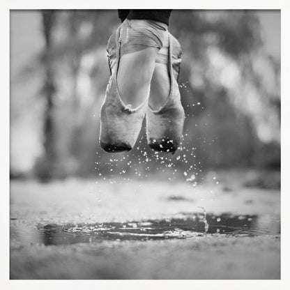 A close-up, black and white photograph of a ballerina's feet in worn pointe shoes, captured in mid-air above a puddle, with water splashing upwards. The motion is frozen, highlighting the contrast between grace and a raw outdoor element. The image is presented in a light wood frame. Poster